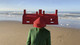 Portrait from 2020 Self-Digitalisation by El Último Grito, taken in Stockholm during their Iaspis residency. A figure stands on a beach wearing a vivid red, house-shaped headpiece with chimney forms and window cut-outs, paired with a structured green coat. The sea and sky create a serene backdrop, contrasting the surreal, architectural wearable.